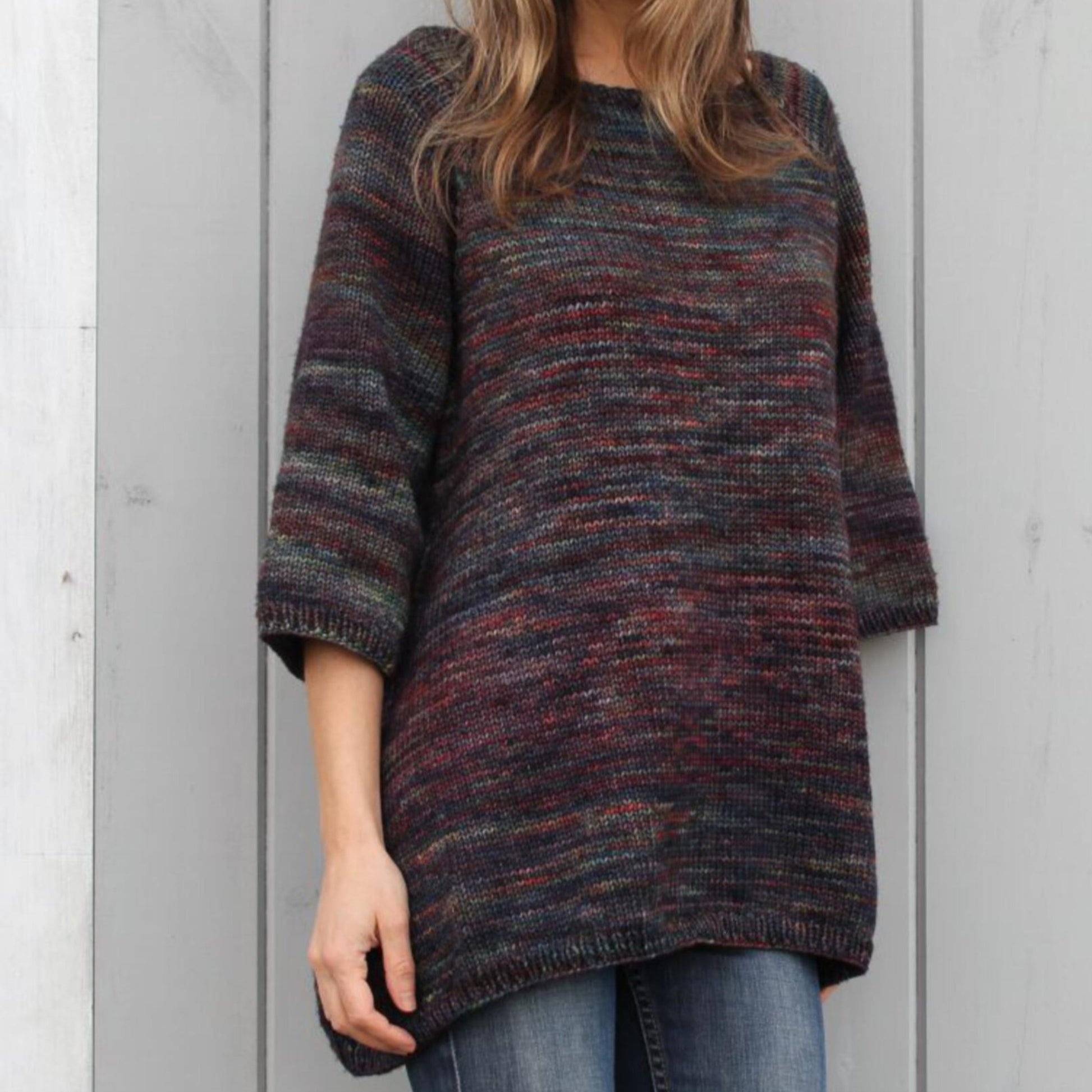 A person is seen standing in front of a gray wooden wall, wearing the Top Down Trapeze Pullover by Bryson Distributing, Inc. This multicolored, loose-fit knitted sweater features 3/4 sleeves and is crafted from variegated yarns. The sweater falls below the hips and is paired perfectly with blue jeans. The individual's face is not visible in the image.