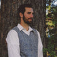 A man with short dark hair and a beard is leaning against a tree trunk. He is wearing a white dress shirt and a printed V-neck vest from Bryson Distributing, Inc.'s mens-vest-v-neck-pattern-printed collection. The background features foliage and trees, indicating an outdoor setting. The man appears to be looking slightly to his left.