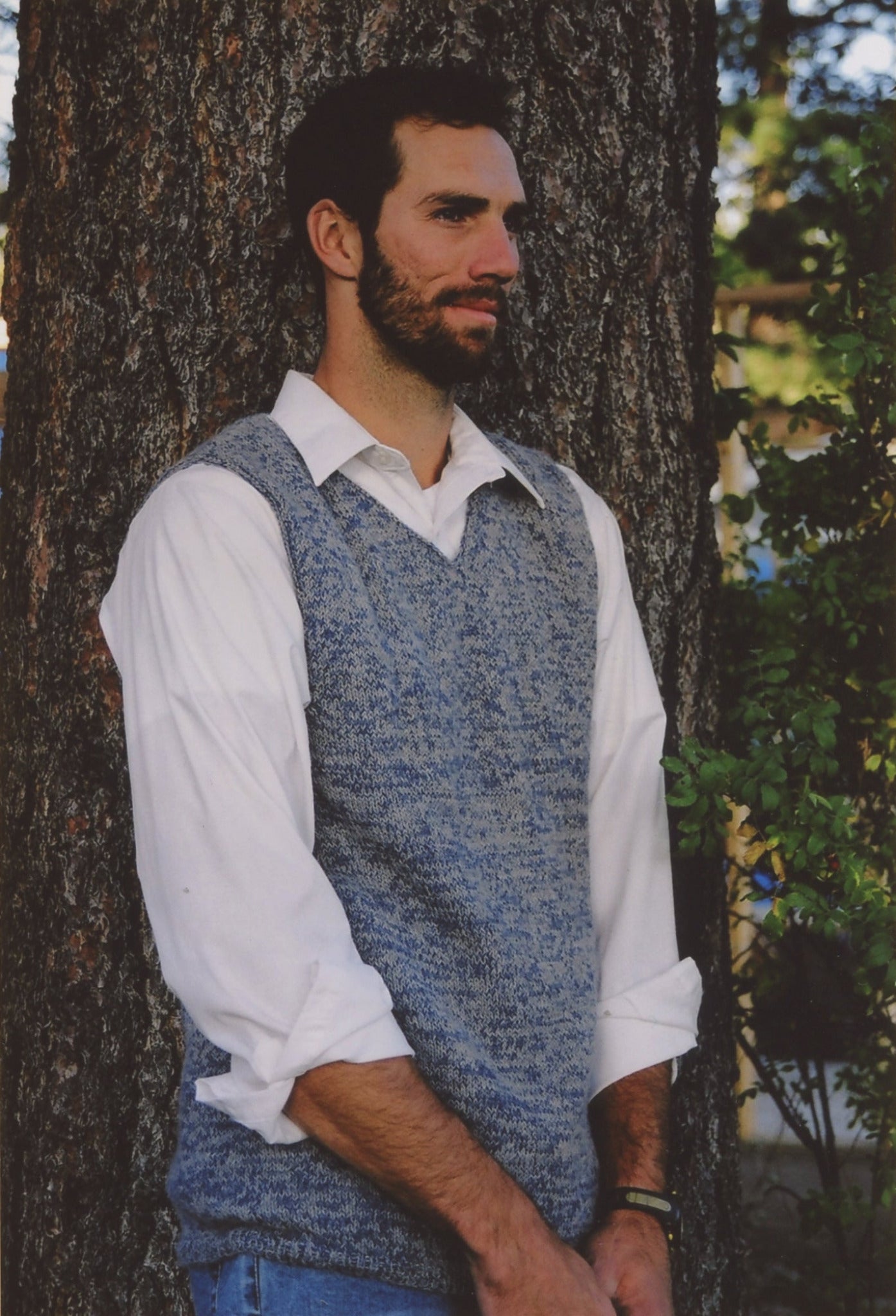 A man with dark hair and a beard stands by a large tree. He is wearing a simple white dress shirt with rolled-up sleeves under a mens-vest-v-neck-pattern-printed by Bryson Distributing, Inc. With a thoughtful expression and his hands clasped in front of him, the design of his outfit complements the green plants visible in the background.