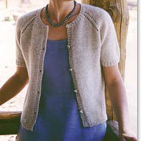 A person wearing Bryson Distributing, Inc.'s light beige Summer Cardigan over a blue dress. This short-sleeved knit cardigan features a round neckline, buttoned front, and eyelet details along the shoulder seams. This versatile pattern is perfect for any occasion. The person is standing near a wooden structure with a blurred background.