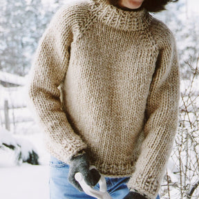 A person wearing the Weekend Neck Down Pullover by Bryson Distributing, Inc., a thick beige raglan sleeve sweater, along with grey gloves, is holding a shovel in a snowy outdoor setting. Only the upper body is visible, with a backdrop of snow-covered trees and bushes.