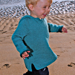 A young child with blonde hair is running on a beach, smiling. The child is wearing Bryson Distributing, Inc.'s aqua-colored Neck Down Children's Hooded Tunic and dark pants. The background shows the sandy beach and water.