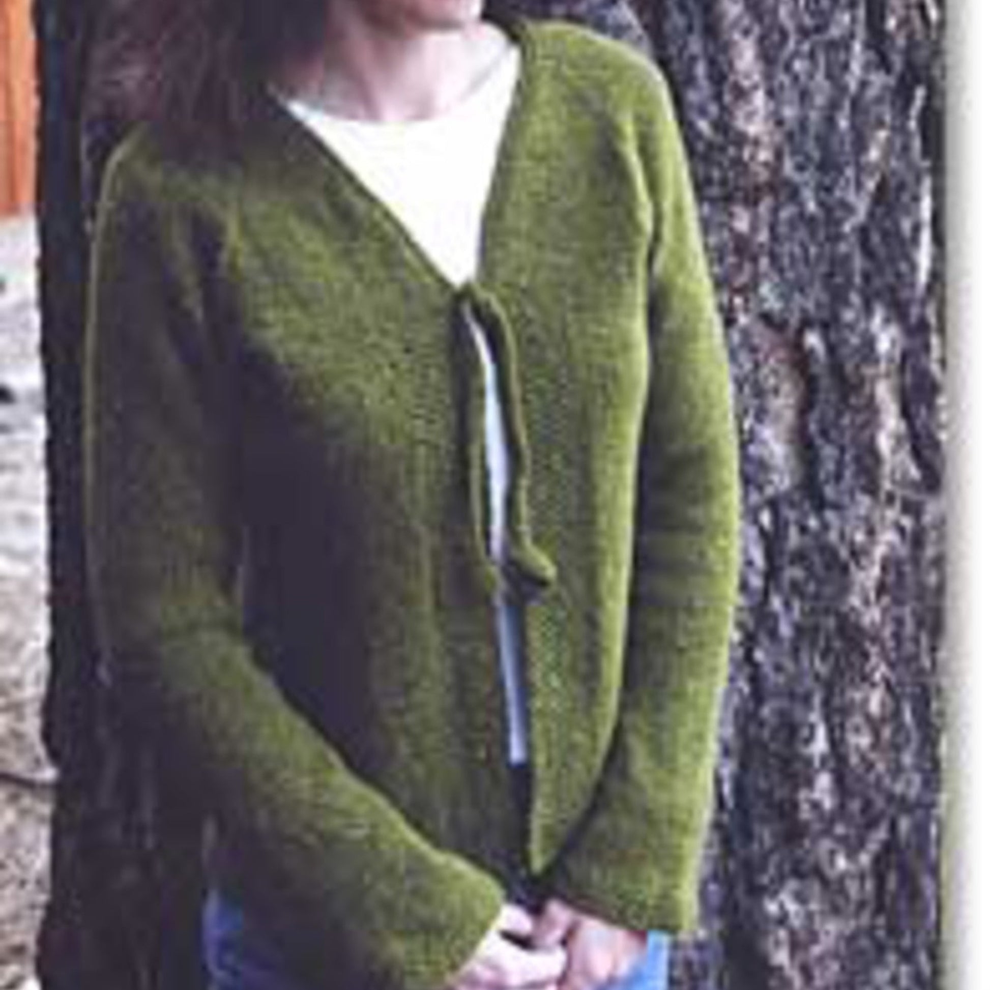 A person standing in front of a tree, partially in view from the chest down, wears Bryson Distributing, Inc.'s Neck Down V-Neck Cardigan with Tie over a white top. The comfortable green cardigan is tied at the front with a knitted bow. The background shows the bark of the tree and the ground beneath it.