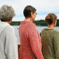 Three people, each wearing a differently colored Very Versatile Neck Down Hooded Pullover by Bryson Distributing, Inc., stand on a wooden deck overlooking a calm body of water with trees in the background. They are facing away from the camera, gazing toward the horizon under a lightly cloudy sky.