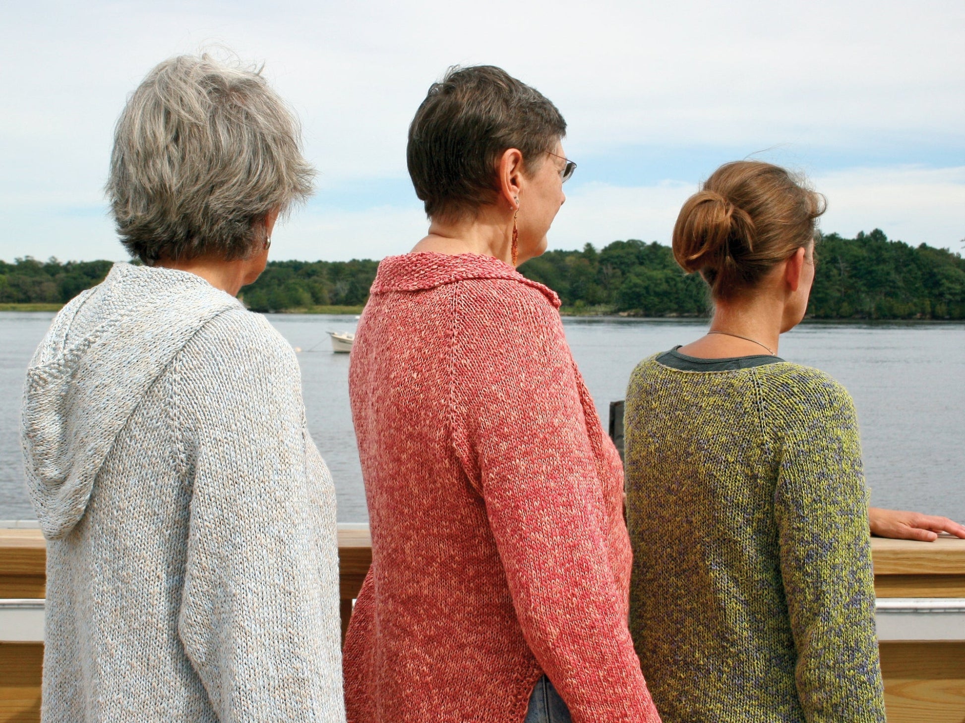 Three people, each wearing a differently colored Very Versatile Neck Down Hooded Pullover by Bryson Distributing, Inc., stand on a wooden deck overlooking a calm body of water with trees in the background. They are facing away from the camera, gazing toward the horizon under a lightly cloudy sky.