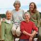 Six women stand and sit together outside, smiling. They wear colorful sweaters in shades of yellow, green, blue, red, and pink. One sports the Very Versatile Neck Down Hooded Pullover from Bryson Distributing, Inc., which offers the perfect fit. Trees and a lake are visible in the background under a clear blue sky. They appear happy and relaxed, enjoying each other's company.