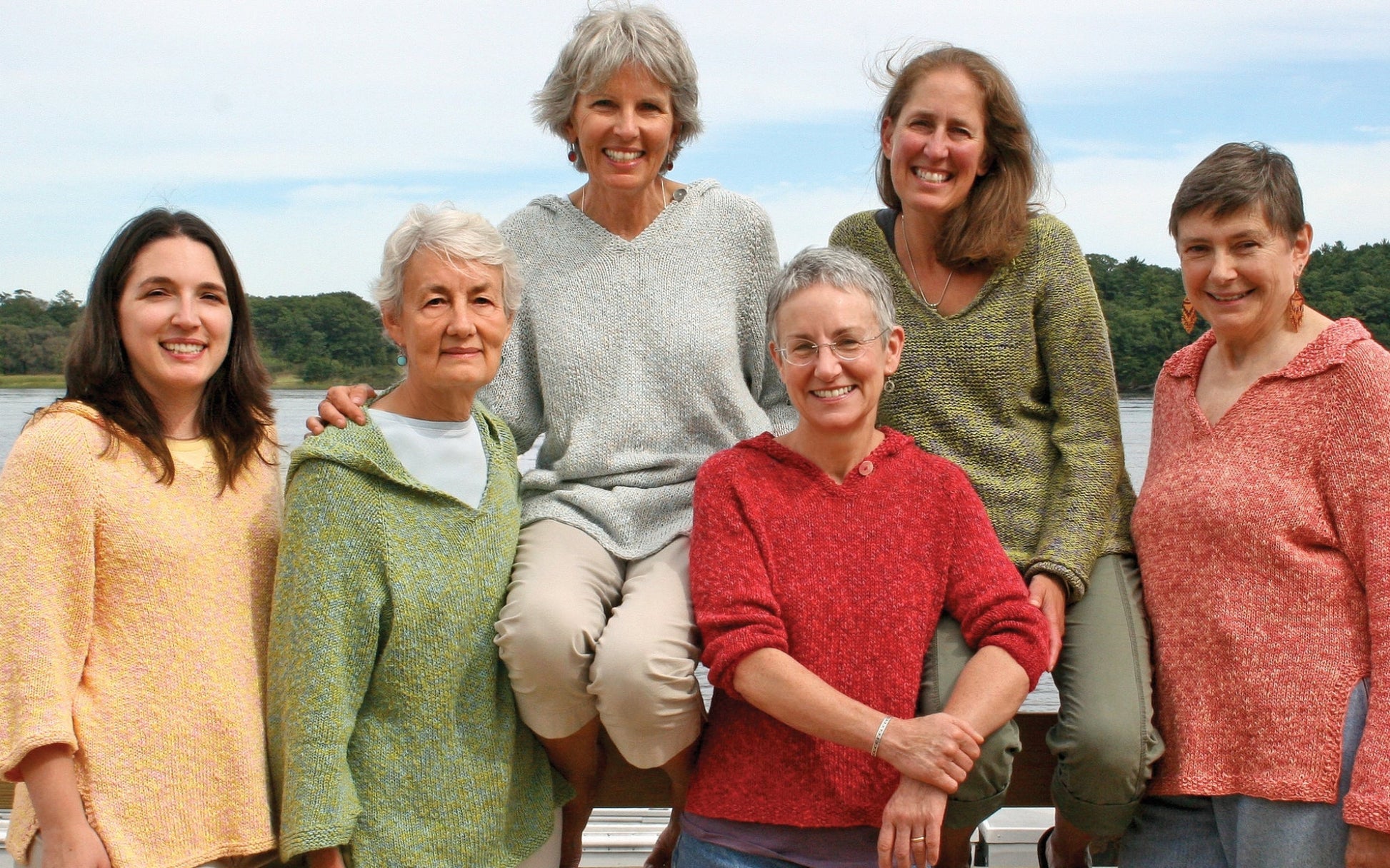 Six women stand and sit together outside, smiling. They wear colorful sweaters in shades of yellow, green, blue, red, and pink. One sports the Very Versatile Neck Down Hooded Pullover from Bryson Distributing, Inc., which offers the perfect fit. Trees and a lake are visible in the background under a clear blue sky. They appear happy and relaxed, enjoying each other's company.