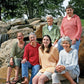 Six women are seated on large rocks in an outdoor setting, with trees and a waterfall in the background. They are all smiling at the camera, wearing different colored Very Versatile Neck Down Hooded Pullovers from Bryson Distributing, Inc., which seem like a perfect fit.