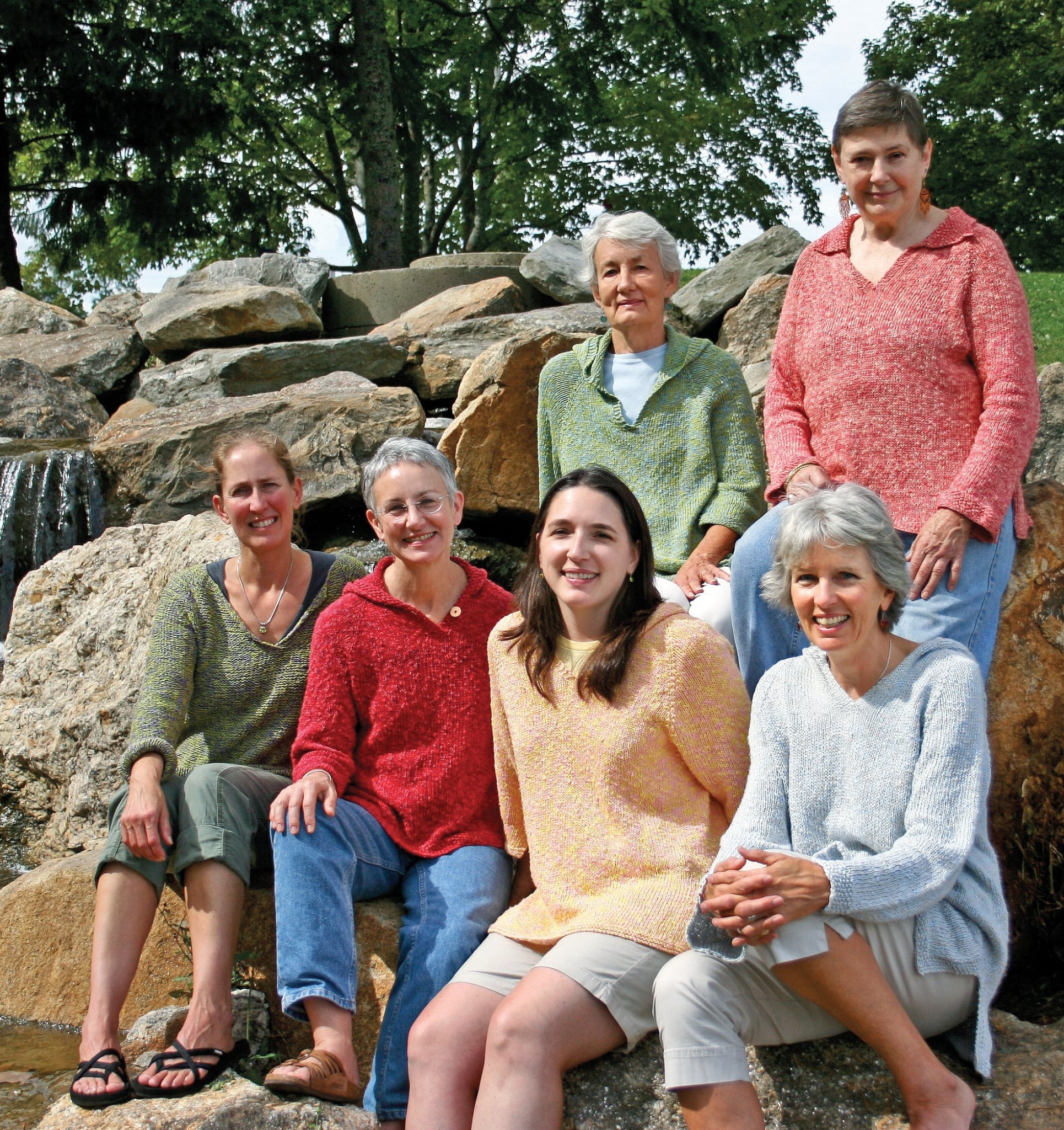 Six women are seated on large rocks in an outdoor setting, with trees and a waterfall in the background. They are all smiling at the camera, wearing different colored Very Versatile Neck Down Hooded Pullovers from Bryson Distributing, Inc., which seem like a perfect fit.