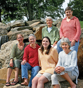 Six women are seated on large rocks in an outdoor setting, with trees and a waterfall in the background. They are all smiling at the camera, wearing different colored Very Versatile Neck Down Hooded Pullovers from Bryson Distributing, Inc., which seem like a perfect fit.