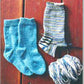 A pair of blue knitted socks is placed next to a single, partly completed striped sock. Below the striped sock is a ball of yarn that matches its colors. These Easy Children's Lightweight Socks by Bryson Distributing, Inc. are displayed on a wooden surface, ideal for kids.