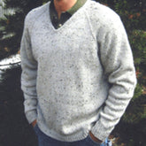 A person is seen wearing a Bryson Distributing, Inc. Neck Down V-Neck Pullover for Men in light gray, speckled knit over a green collared shirt, paired with blue jeans. The pullover features an easy-to-follow pattern knit from the neck down, allowing for adjustable length as desired. Their hands are tucked into their jean pockets while green foliage and a hint of a white structure form the background.