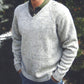 A person is seen wearing a Bryson Distributing, Inc. Neck Down V-Neck Pullover for Men in light gray, speckled knit over a green collared shirt, paired with blue jeans. The pullover features an easy-to-follow pattern knit from the neck down, allowing for adjustable length as desired. Their hands are tucked into their jean pockets while green foliage and a hint of a white structure form the background.