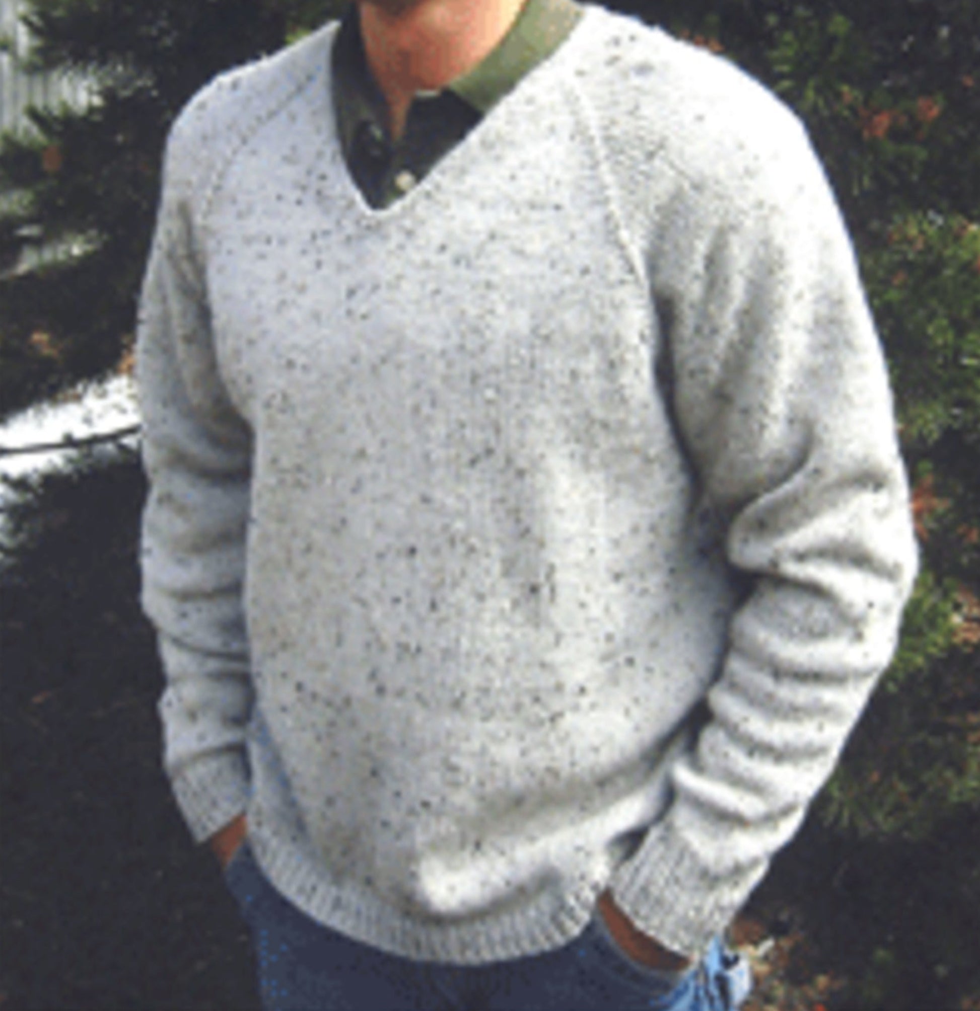 A person is seen wearing a Bryson Distributing, Inc. Neck Down V-Neck Pullover for Men in light gray, speckled knit over a green collared shirt, paired with blue jeans. The pullover features an easy-to-follow pattern knit from the neck down, allowing for adjustable length as desired. Their hands are tucked into their jean pockets while green foliage and a hint of a white structure form the background.