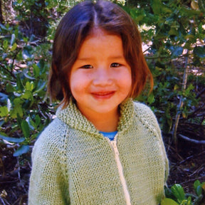 A young child with shoulder-length brown hair and a green Children's Bulky Neck Down Jacket by Bryson Distributing, Inc. stands outdoors, smiling at the camera. The background is filled with lush green foliage and sunlight filtering through the leaves. The cozy jacket highlights the charm of childhood in nature.