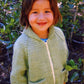 A young child with shoulder-length hair smiles at the camera while standing outdoors. They are wearing the Children's Bulky Neck Down Jacket by Bryson Distributing, Inc., a green knitted sweater with a front zipper and two pockets, featuring a cozy shawl collar. The background is filled with lush green foliage.