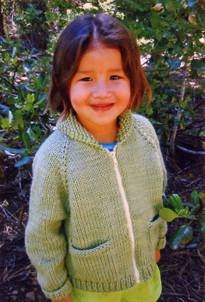 A young child with shoulder-length hair smiles at the camera while standing outdoors. They are wearing the Children's Bulky Neck Down Jacket by Bryson Distributing, Inc., a green knitted sweater with a front zipper and two pockets, featuring a cozy shawl collar. The background is filled with lush green foliage.