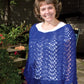 A person stands outside in a garden, smiling at the camera. They are wearing a white shirt underneath Bryson Distributing, Inc.'s Easy Lace Poncho in blue and blue jeans. The background includes greenery, flowers, and partial shadows from trees or plants.