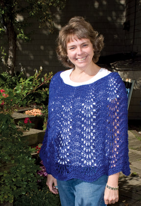 A person stands outside in a garden, smiling at the camera. They are wearing a white shirt underneath Bryson Distributing, Inc.'s Easy Lace Poncho in blue and blue jeans. The background includes greenery, flowers, and partial shadows from trees or plants.