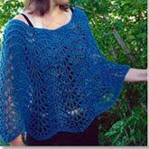 A person donning Bryson Distributing, Inc.'s Easy Lace Poncho, which boasts a loose-knit blue crochet design with a fan-like pattern, stands against a backdrop of lush green foliage. This stylish poncho drapes comfortably over the person's shoulders and upper arms.