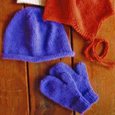 A Bryson Distributing, Inc. Basic Hat & Mitten Set for Children, featuring a purple stockinette stitch hat and matching purple mittens, is displayed on a wooden surface. In the background, an orange bonnet with ties and part of a cream-colored knitted item are also visible, showcasing different styles.