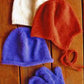 Three knitted hats and a pair of mittens from Bryson Distributing, Inc.'s Basic Hat & Mitten Set for Children are displayed on a wooden surface. The hats come in white, red, and blue. The red hat features earflaps and strings, while the blue stockinette stitch hat is paired with matching blue mittens.