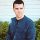 A man in a cozy Men's Neck Down Henley Knitting from Bryson Distributing, Inc. and a dark undershirt is standing in front of a house with white siding. He gazes to the side with a neutral expression, his short, dark hair perfectly styled. Green plants flourish near the base of the house, adding a touch of nature to the scene.