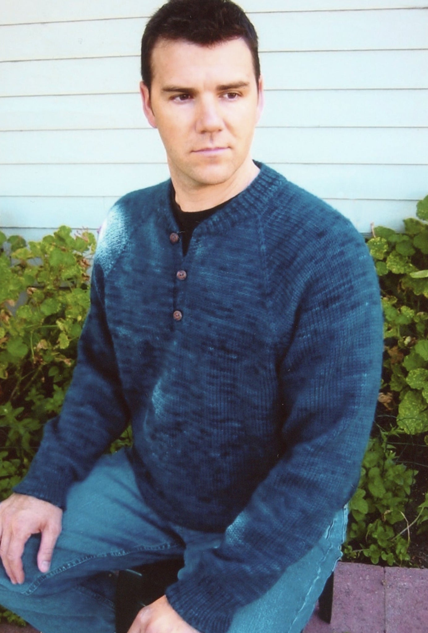 A man sporting the Men's Neck Down Henley Knitting by Bryson Distributing, Inc. and a pair of jeans is seated on a bench in front of a light gray wooden wall, with vibrant green plants in the background. He has short dark hair and is gazing slightly to the side.