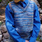 A child stands outdoors near a rocky background, wearing a blue long-sleeve shirt paired with Bryson Distributing, Inc.'s Basic Vest for Children, which is knitted in blue and multicolors. The child has their hands in their pockets. Only the upper body is visible.