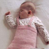 A baby sleeps peacefully on a white surface, wearing a white long-sleeve shirt adorned with small patterns and a knitted pink sleeveless garment. Dressed in cozy Baby Bottoms from Bryson Distributing, Inc., they lie on their side with one arm bent upward and the other resting by their side.