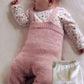 A baby wearing a white long-sleeve shirt with small patterns and pink knitted overalls is sleeping on a white surface. Next to the baby is a pair of Baby Bottoms from Bryson Distributing, Inc., featuring an elastic waist. The baby is sleeping on its back with one arm slightly raised.