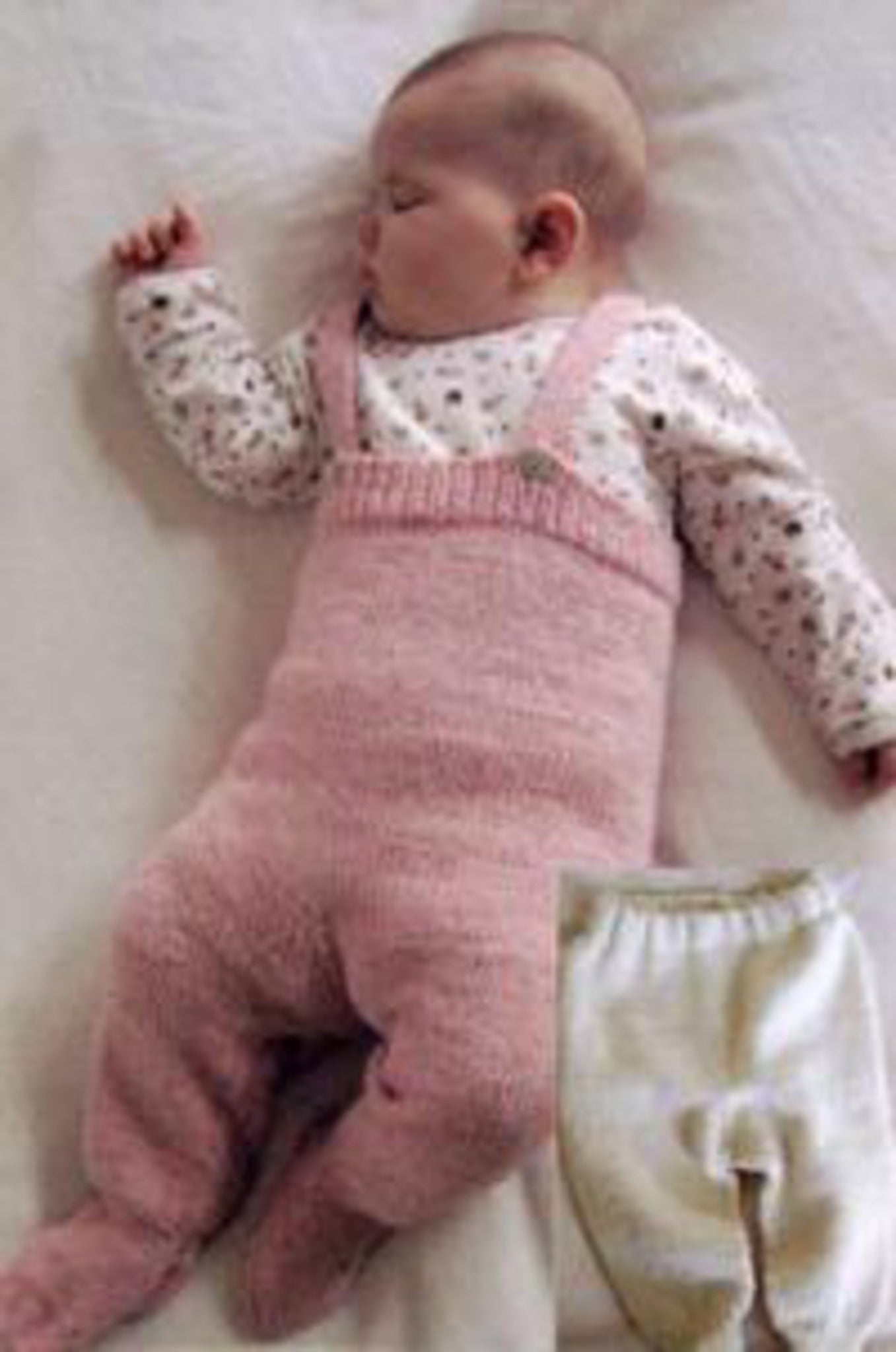 A baby wearing a white long-sleeve shirt with small patterns and pink knitted overalls is sleeping on a white surface. Next to the baby is a pair of Baby Bottoms from Bryson Distributing, Inc., featuring an elastic waist. The baby is sleeping on its back with one arm slightly raised.