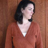 A woman with shoulder-length dark hair is standing against a wooden wall. She is wearing a rust-colored Neckdown Wrap Cardigan by Bryson Distributing, Inc., over a white top, and she is looking to the right with a neutral expression. The background consists of vertical wooden panels.