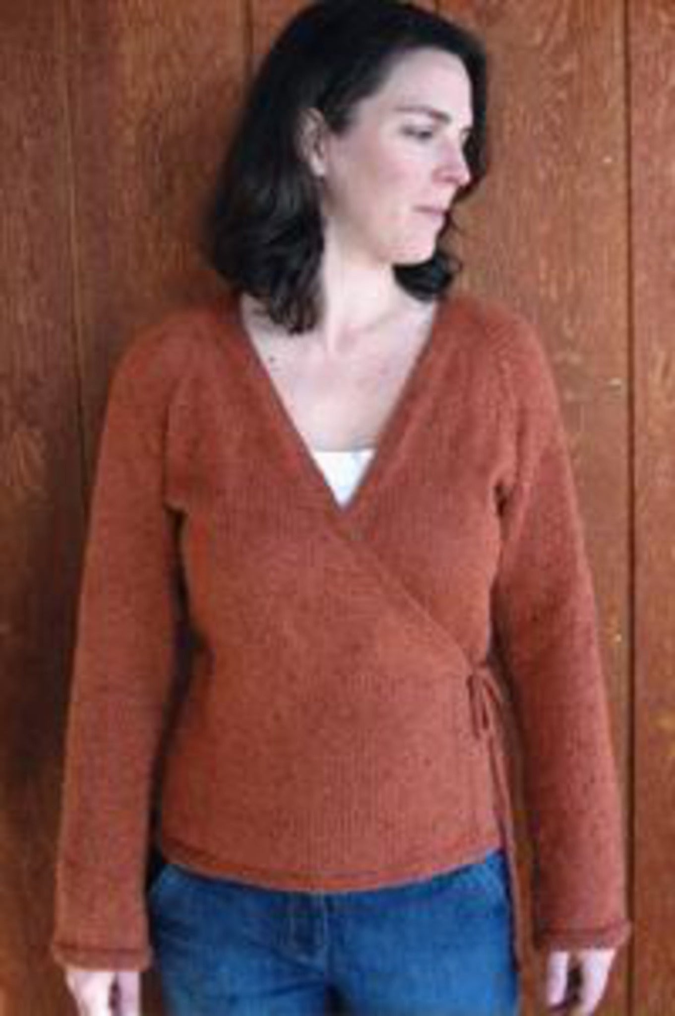 A woman with shoulder-length brown hair stands in front of a wooden wall, wearing the rust-colored Neckdown Wrap Cardigan by Bryson Distributing, Inc. and blue jeans. She is looking away from the camera.