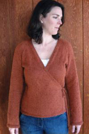 A woman with shoulder-length brown hair stands in front of a wooden wall, wearing the rust-colored Neckdown Wrap Cardigan by Bryson Distributing, Inc. and blue jeans. She is looking away from the camera.