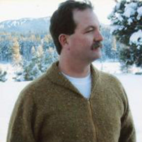 A person wearing the Neckdown Cardigan for Men by Bryson Distributing, Inc., stands in a snowy outdoor setting, with trees and snow-covered mountains in the background. The individual is looking to the side, and the landscape indicates a cold, winter environment.