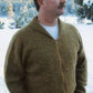 A mustached man sporting a brown Neckdown Cardigan for Men by Bryson Distributing, Inc., with a white shirt underneath, stands outdoors with his hands in his pockets. The background features a snowy landscape with trees and mountains.