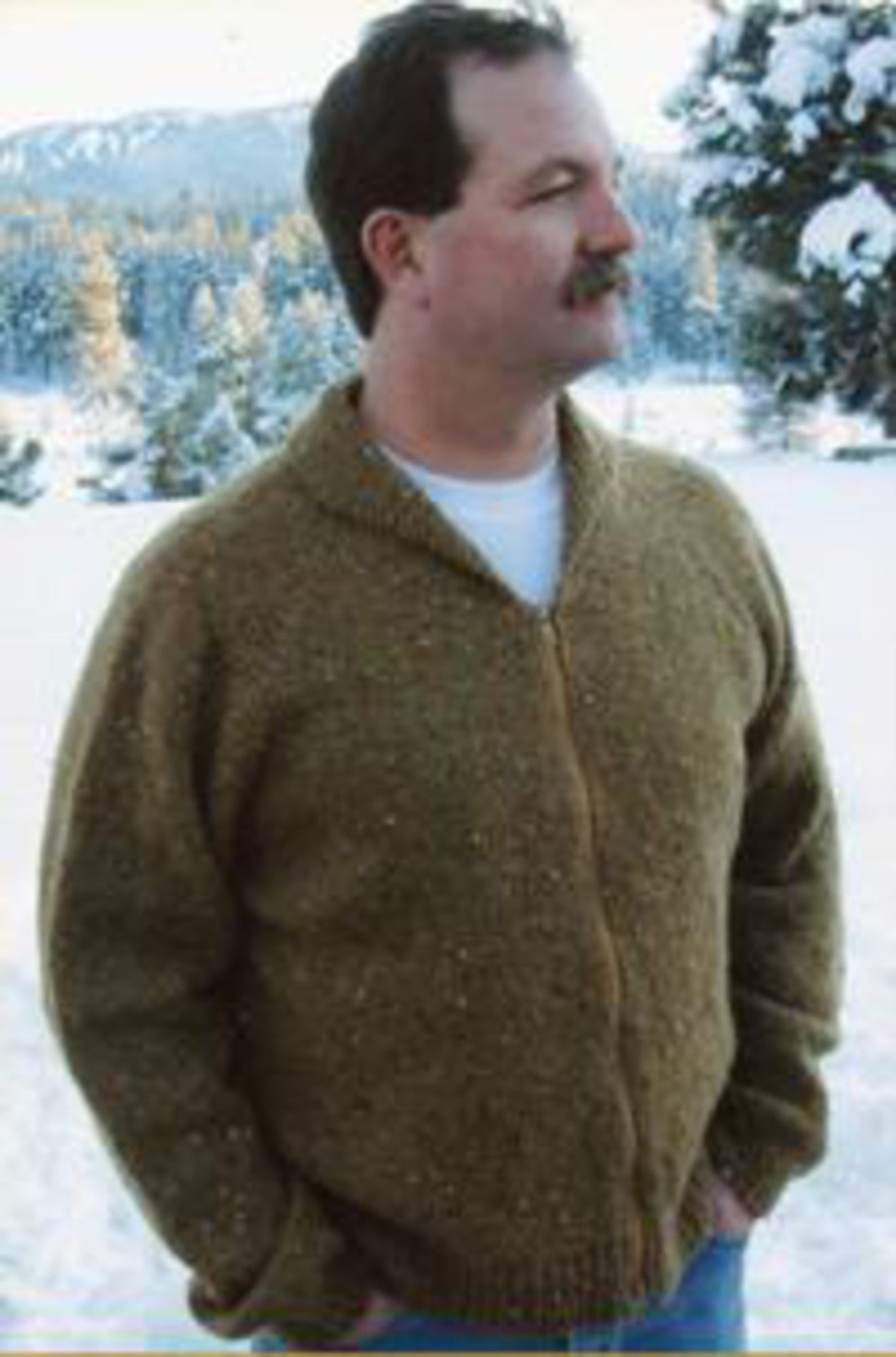 A mustached man sporting a brown Neckdown Cardigan for Men by Bryson Distributing, Inc., with a white shirt underneath, stands outdoors with his hands in his pockets. The background features a snowy landscape with trees and mountains.