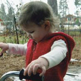 A young child with short hair is outdoors, wearing a cozy Bryson Distributing, Inc. Bulky Hooded Vest for Children over a white shirt. The child is holding onto the handles of a walking aid, concentrating on their activity. Trees and some blurred background elements are visible behind them.