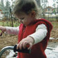 A toddler wearing a Bryson Distributing, Inc. Bulky Hooded Vest for Children in red and a white long-sleeve shirt is holding onto a tricycle handlebar outdoors. The ground is covered with dry pine needles and patches of snow. The background includes trees and a house at a distance.