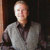 A middle-aged man with light hair stands smiling in a doorway. He is wearing the Basic Cardigan Vest for Men by Bryson Distributing, Inc., layered over a dark brown button-up shirt. The background is relatively dark, making the man the focal point of the image.