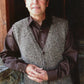 A man with light brown hair stands leaning against a rustic wooden wall. He is wearing a brown long-sleeve shirt and Bryson Distributing, Inc.'s gray, speckled Basic Cardigan Vest for Men. He gazes upwards with a slight smile, his hands gently clasped in front of him.