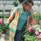 A woman with brown hair tied back in a ponytail is wearing the Neck Down Swing Coat, a brightly colored, striped cardigan coat made of worsted weight yarn from Bryson Distributing, Inc. She is in a greenhouse, surrounded by pots of pink flowers, examining one with her hand. She is also wearing a blue shirt and jeans.