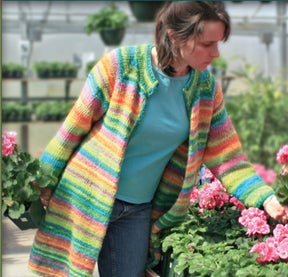 A woman with brown hair tied back in a ponytail is wearing the Neck Down Swing Coat, a brightly colored, striped cardigan coat made of worsted weight yarn from Bryson Distributing, Inc. She is in a greenhouse, surrounded by pots of pink flowers, examining one with her hand. She is also wearing a blue shirt and jeans.