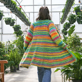 A person with long hair, seen from behind, walks in a greenhouse wearing the brightly colored Neck Down Swing Coat by Bryson Distributing, Inc., made with worsted weight yarn. The greenhouse is abundant with lush green plants, creating a vibrant and lively atmosphere.