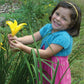 A young girl wearing Bryson Distributing, Inc.'s Little Girl's Shrug in blue over a pink sundress smiles brightly while holding a yellow flower. She accessorizes with a yellow headband and stands amidst green foliage in a garden, looking completely comfortable and at ease.