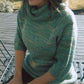 A person seated outdoors on a wooden deck in a chair with a slight smile, wearing the stylish Neck Down Cowl Collar Pullover by Bryson Distributing, Inc. The sunlight highlights the multicolored yarn of the comfortable greenish-blue knit sweater that features a cowl neck and three-quarter sleeves. Their head is partly out of the frame.