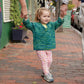 A cheerful young girl with blonde hair tied in a ponytail walks on a brick sidewalk, holding hands with two adults, one on each side of her. She's wearing a lightweight Children's V-Neck Neck Down Pullover by Bryson Distributing, Inc., paired with floral pants. The background features a green wall and a potted plant.