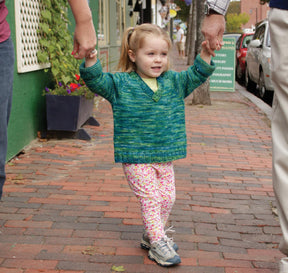 A cheerful young girl with blonde hair tied in a ponytail walks on a brick sidewalk, holding hands with two adults, one on each side of her. She's wearing a lightweight Children's V-Neck Neck Down Pullover by Bryson Distributing, Inc., paired with floral pants. The background features a green wall and a potted plant.