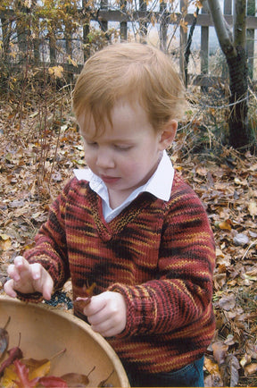 A young child with ginger hair stands outdoors holding a bowl filled with autumn leaves. The child is wearing the Children's V-Neck, Neck Down Pullover by Bryson Distributing, Inc., over a white collared shirt and is surrounded by fallen leaves and a wooden fence in the background.